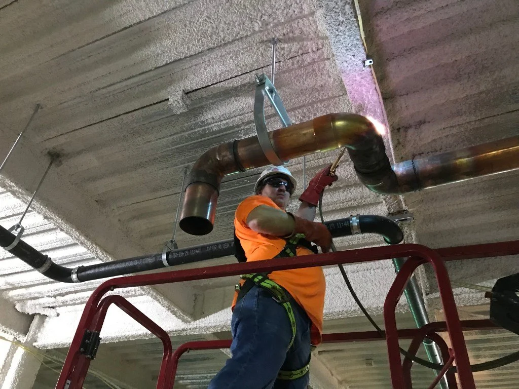 Dan Hogan soldering copper pipe on a construction site while wearing a hard hat and safety harness